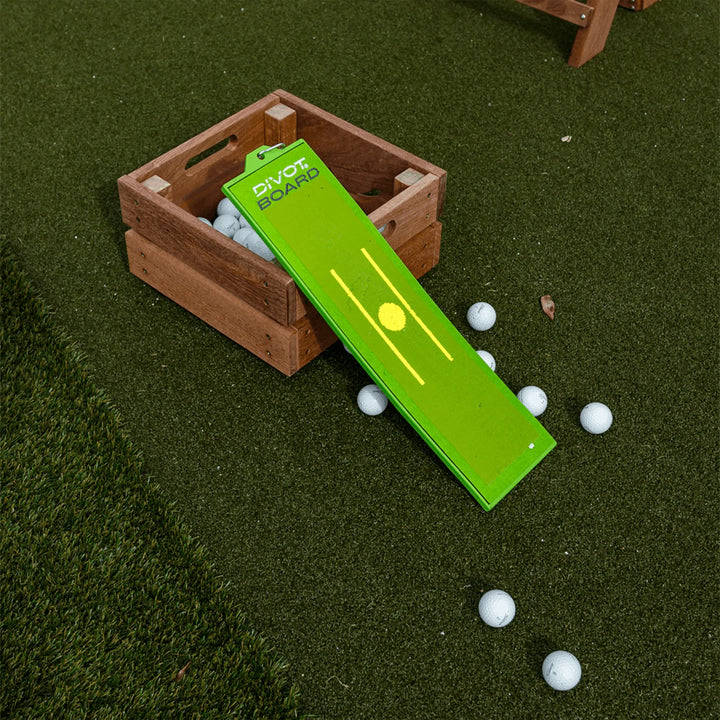Divot Board propped against a wooden crate of range balls on turf, with balls scattered across the grass