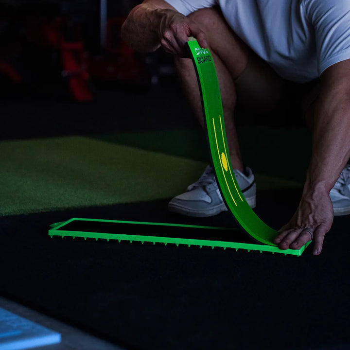 Peeling the used Divot Board pad off the green board frame, left empty on a black mat in an indoor hitting bay