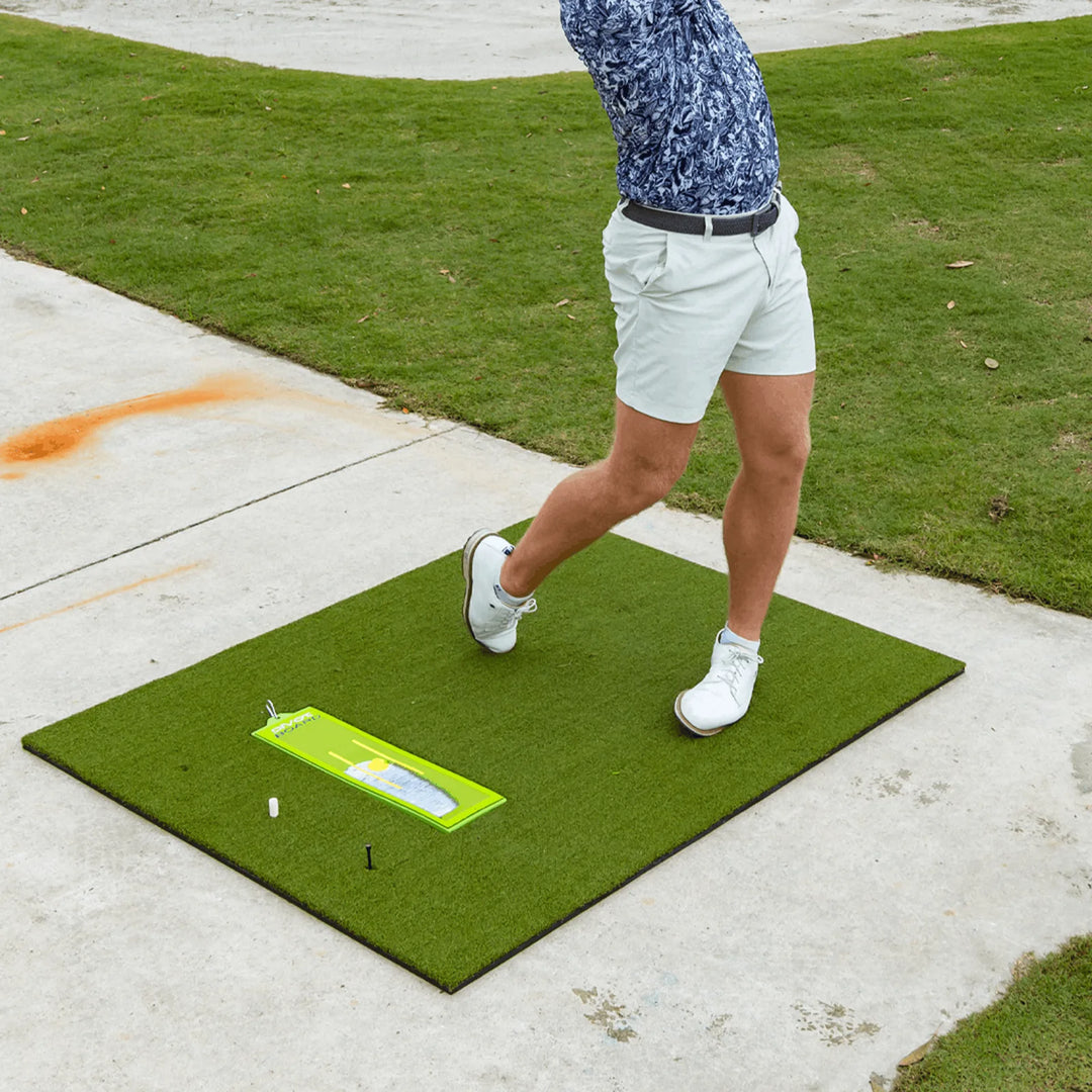 Golfer in follow-through on the Divot Board Turf Mat, with the Divot Board on the turf showing a strike mark