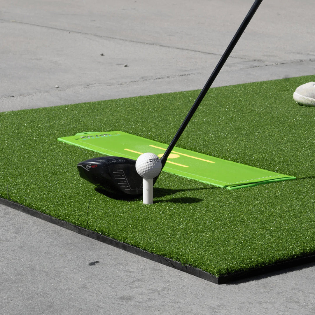 Driver at address behind a teed golf ball on the turf hitting mat, Divot Board lying flat alongside