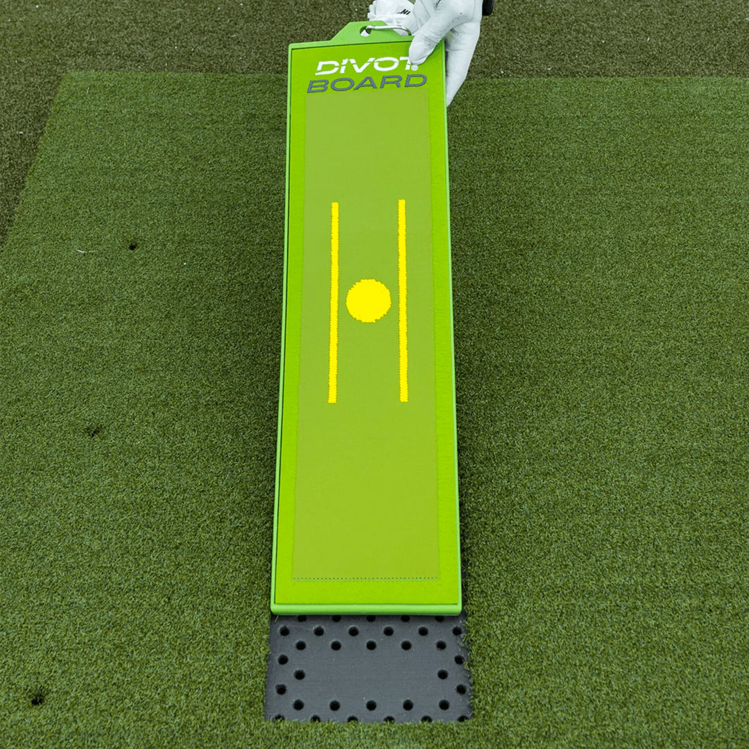 Divot Board face up on the turf mat, yellow alignment lines centered, gloved hand at top, grey foam pad below